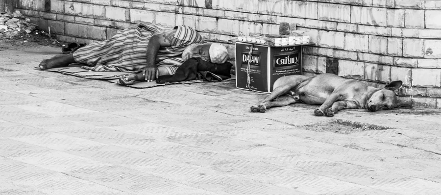 Two people sleeping beside a dog on a pavement in a black and white photograph.